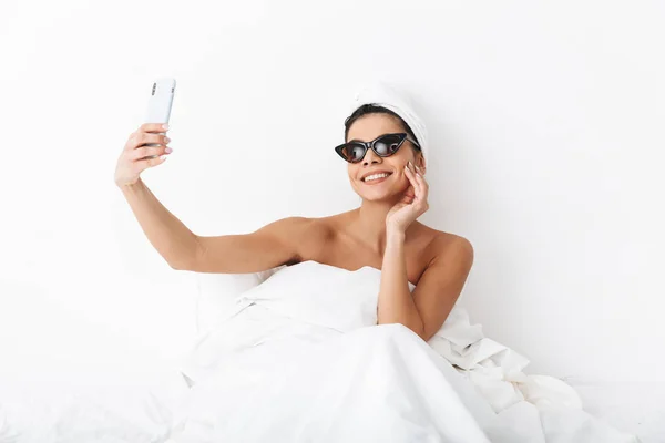 Όμορφη συναισθηματική γυναίκα με πετσέτα στο κεφάλι βρίσκεται στο κρεβάτι κάτω από την κουβέρτα απομονωμένη πάνω από λευκό τοίχο φόντο φορώντας γυαλιά ηλίου πάρει μια selfie από το κινητό τηλέφωνο. — Φωτογραφία Αρχείου
