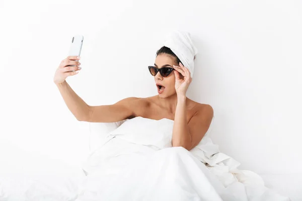 Hermosa mujer emocional con toalla en la cabeza se encuentra en la cama debajo de la manta aislada sobre fondo blanco de la pared con gafas de sol tomar una selfie por teléfono móvil . — Foto de Stock