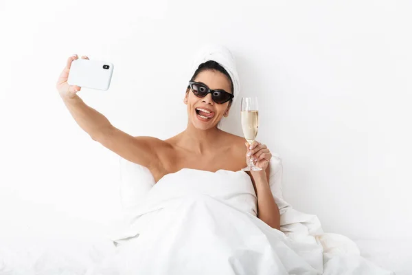 Bella donna emotiva con asciugamano sulla testa si trova nel letto sotto coperta isolata su sfondo bianco parete indossando occhiali da sole prendere un selfie dal telefono cellulare . — Foto Stock
