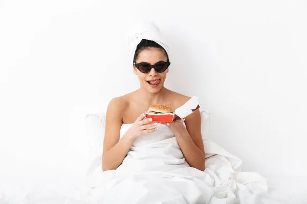 Mujer emocional con toalla en la cabeza se encuentra en la cama bajo una manta aislada sobre fondo blanco de la pared con gafas de sol comer hamburguesa . — Foto de Stock