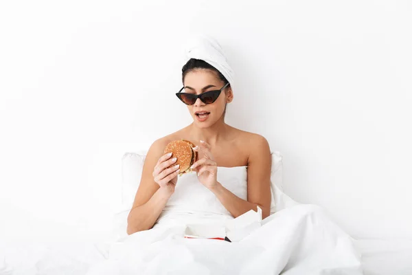 Mulher emocional com toalha na cabeça encontra-se na cama sob cobertor isolado sobre fundo da parede branca usando óculos de sol comer hambúrguer . — Fotografia de Stock