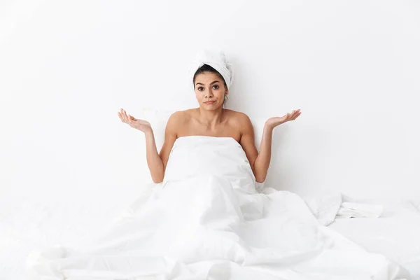 Vackra förvirrad fantastisk kvinna med handduk på huvudet ligger i sängen under filt isolerad över vit vägg bakgrund. — Stockfoto