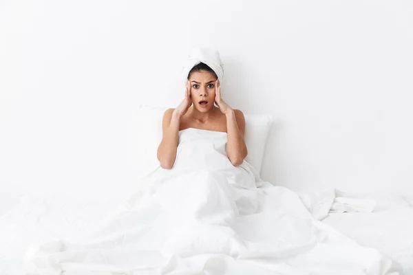 Geschokt emotionele verbazingwekkende vrouw met handdoek op hoofd ligt in bed onder deken geïsoleerd over witte muur achtergrond. — Stockfoto