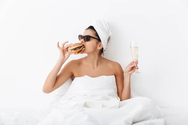 Donna emotiva con asciugamano sulla testa si trova a letto sotto coperta isolato su sfondo bianco parete indossando occhiali da sole mangiare hamburger . — Foto Stock