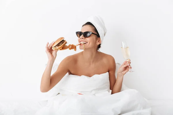 Donna emotiva con asciugamano sulla testa si trova a letto sotto coperta isolato su sfondo bianco parete indossando occhiali da sole mangiare hamburger . — Foto Stock