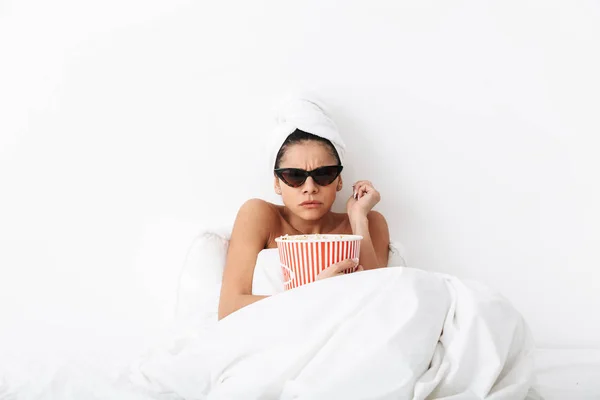 Mujer asustada conmocionada con toalla en la cabeza se encuentra en la cama debajo de una manta aislada sobre fondo blanco de la pared con gafas de sol comer palomitas de maíz ver película tv . —  Fotos de Stock