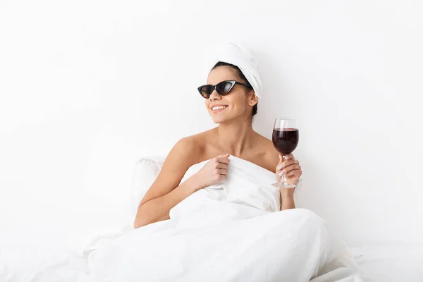 Donna con asciugamano sulla testa si trova nel letto sotto coperta isolato su sfondo bianco parete indossando occhiali da sole bere vino . — Foto Stock