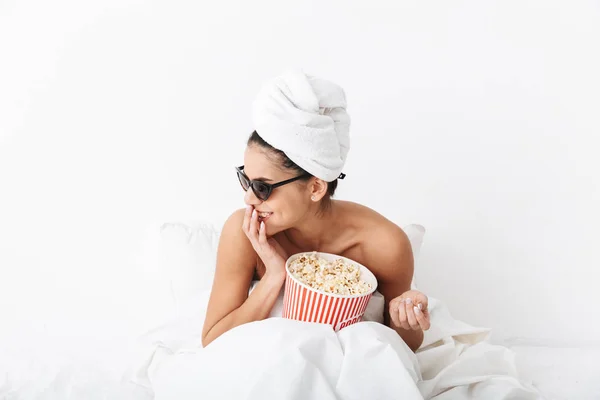 Lachende Frau mit Handtuch auf dem Kopf liegt im Bett unter Decke isoliert über weißem Wandhintergrund mit Sonnenbrille Popcorn essen Filmfernsehen. — Stockfoto