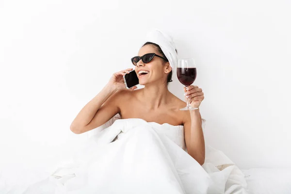 Piękna kobieta z ręcznikiem na głowie leży w łóżku pod kocem na białym tle przez białą ścianę w okularach do picia, rozmawiając przez telefon komórkowy. — Zdjęcie stockowe