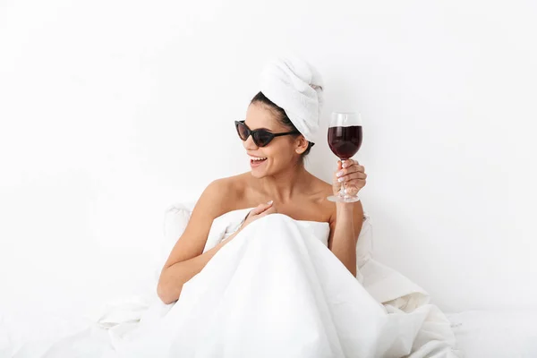 Mujer con toalla en la cabeza se encuentra en la cama bajo una manta aislada sobre fondo blanco de la pared con gafas de sol beber vino . — Foto de Stock