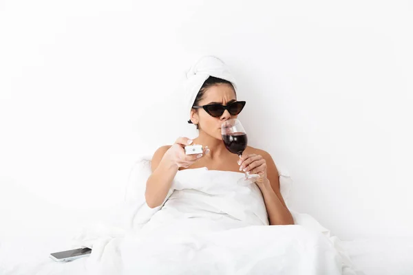 Donna con asciugamano sulla testa si trova nel letto sotto coperta isolato su sfondo bianco parete indossando occhiali da sole bere vino guardare film tv . — Foto Stock