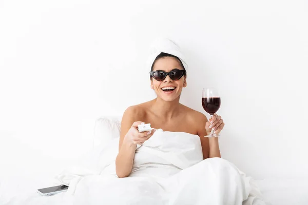 Femme avec serviette sur la tête se trouve dans le lit sous la couverture isolé sur fond de mur blanc portant des lunettes de soleil boire du vin regarder la télévision film . — Photo