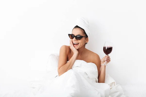 Ridendo donna felice con asciugamano sulla testa si trova nel letto sotto coperta isolato su sfondo bianco parete indossando occhiali da sole bere vino . — Foto Stock