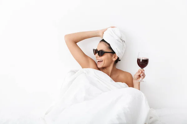 Όμορφο γέλιο χαρούμενη γυναίκα με πετσέτα στο κεφάλι βρίσκεται στο κρεβάτι κάτω από κουβέρτα απομονωμένη πάνω από λευκό τοίχο φόντο φορώντας γυαλιά ηλίου πόσιμο κρασί. — Φωτογραφία Αρχείου