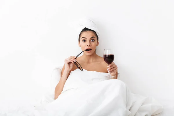 Donna con asciugamano sulla testa si trova nel letto sotto coperta isolata su sfondo bianco parete che tiene occhiali da sole bere vino . — Foto Stock