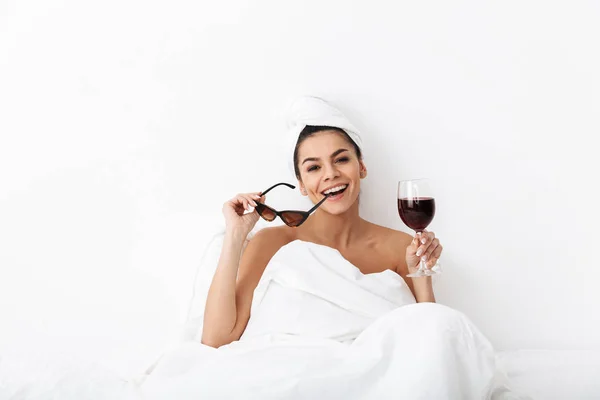 Ευτυχισμένη γυναίκα με πετσέτα στο κεφάλι βρίσκεται στο κρεβάτι κάτω από κουβέρτα απομονωμένη πάνω από λευκό τοίχο φόντο κρατώντας γυαλιά ηλίου πόσιμο κρασί. — Φωτογραφία Αρχείου