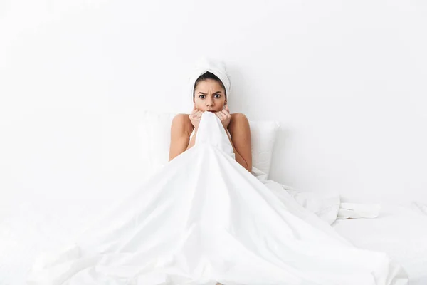 Bang geweldige vrouw met handdoek op hoofd ligt in bed bedekt zichzelf onder deken geïsoleerd over witte muur achtergrond. — Stockfoto