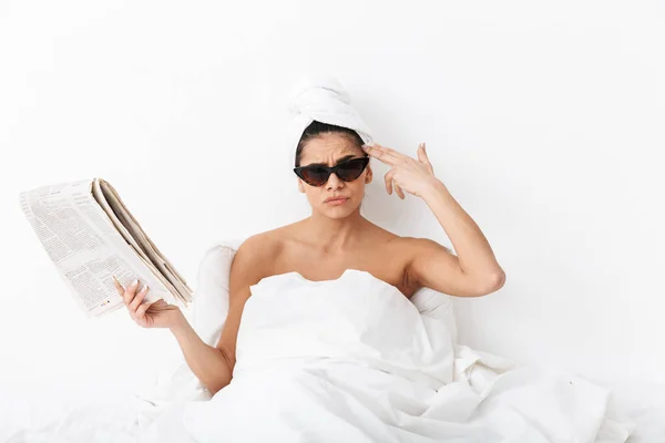 Hermosa mujer con toalla en la cabeza se encuentra en la cama debajo de la manta aislada sobre fondo de pared blanca con gafas de sol leer el periódico . —  Fotos de Stock