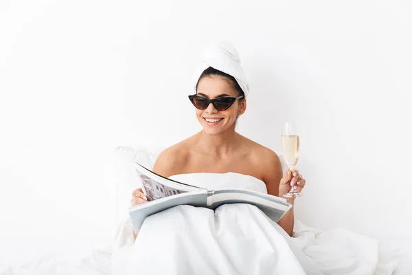 Donna con asciugamano sulla testa si trova nel letto sotto coperta isolato su sfondo bianco parete indossare occhiali da sole leggere rivista . — Foto Stock