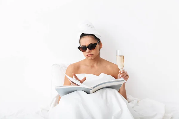 Mujer con toalla en la cabeza se encuentra en la cama debajo de la manta aislada sobre fondo de pared blanca con gafas de sol leer revista . — Foto de Stock