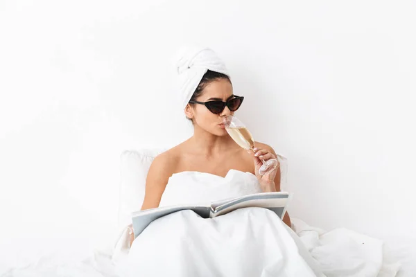 Mujer con toalla en la cabeza se encuentra en la cama debajo de la manta aislada sobre fondo de pared blanca con gafas de sol leer revista . — Foto de Stock