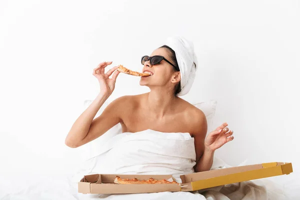 Nő törülköző a fején fekszik az ágyban takaró alatt elszigetelt fehér fal háttér visel napszemüveg eszik pizza. — Stock Fotó
