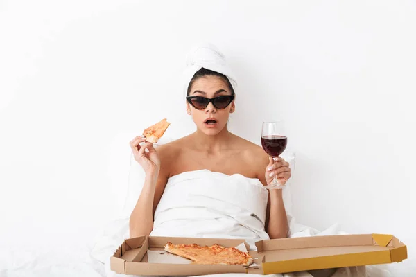 Donna con asciugamano sulla testa si trova nel letto sotto coperta isolato su sfondo bianco parete indossando occhiali da sole mangiare pizza . — Foto Stock