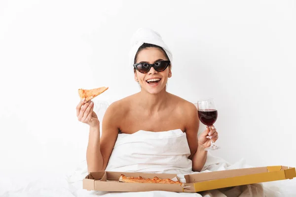 Donna con asciugamano sulla testa si trova nel letto sotto coperta isolato su sfondo bianco parete indossando occhiali da sole mangiare pizza . — Foto Stock