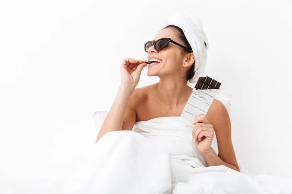 Woman with towel on head lies in bed under blanket isolated over white wall background wearing sunglasses eat chocolate. — 스톡 사진