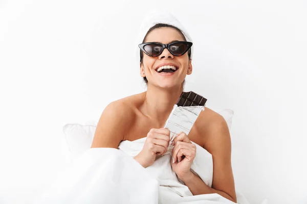 Donna con asciugamano sulla testa si trova nel letto sotto coperta isolato su sfondo bianco parete indossando occhiali da sole mangiare cioccolato . — Foto Stock