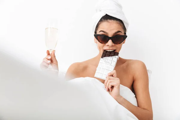 Жінка з рушником на голові лежить в ліжку під ковдрою, ізольована на білому тлі стіни в сонцезахисних окулярах їсть шоколад . — стокове фото