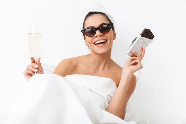 Mujer con toalla en la cabeza se encuentra en la cama bajo una manta aislada sobre fondo blanco de la pared con gafas de sol comer chocolate . —  Fotos de Stock