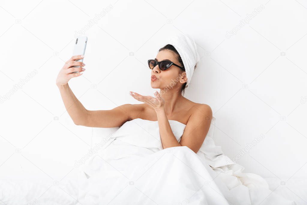 Beautiful emotional woman with towel on head lies in bed under blanket isolated over white wall background wearing sunglasses take a selfie by mobile phone.