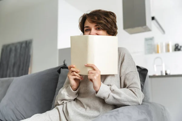 Lächelnde brünette Frau versteckt sich hinter Buch und schaut in die Kamera — Stockfoto