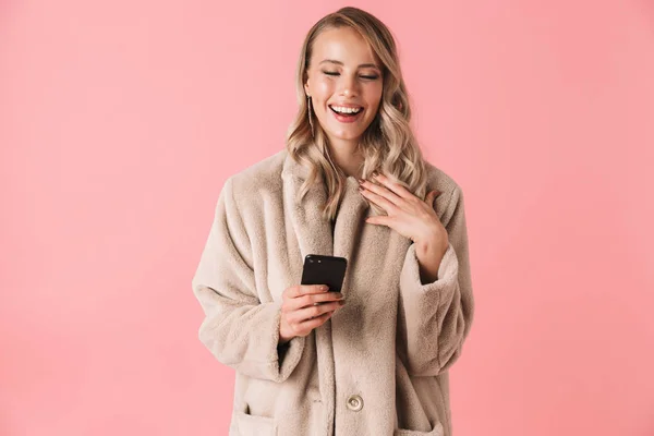 Mujer rubia sonriente usando abrigo de piel sosteniendo teléfono inteligente — Foto de Stock