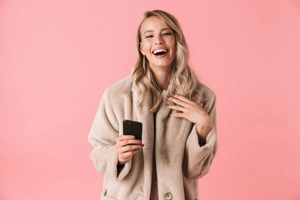 Mujer rubia sonriente usando abrigo de piel sosteniendo teléfono inteligente — Foto de Stock