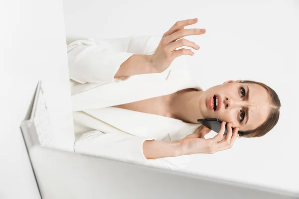 Mulher descontente sentado isolado sobre fundo da parede branca usando computador pc falando por telefone móvel . — Fotografia de Stock