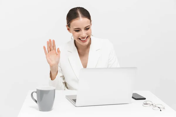 Mulher feliz posando isolado sobre fundo da parede branca usando computador portátil acenando . — Fotografia de Stock