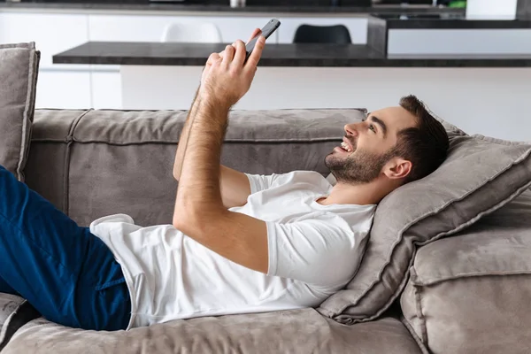 Foto de un chico satisfecho sosteniendo y usando un teléfono inteligente mientras está acostado — Foto de Stock