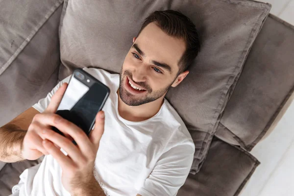 Foto de un chico atractivo sosteniendo y usando un teléfono inteligente mientras miente —  Fotos de Stock