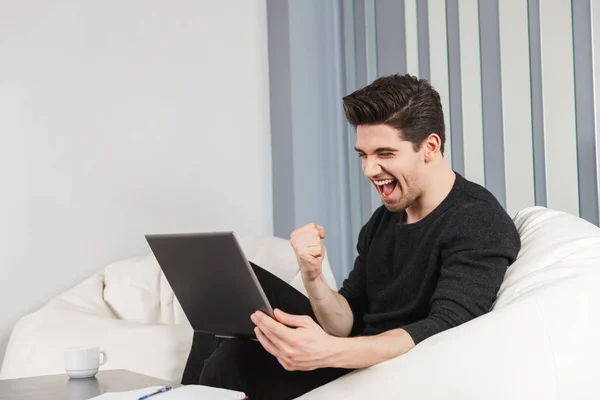 Jovem bonito feliz em casa dentro de casa usando computador portátil fazer gesto vencedor . — Fotografia de Stock