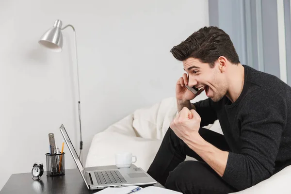 Hombre joven guapo en casa en interiores usando ordenador portátil hablando por teléfono móvil . — Foto de Stock