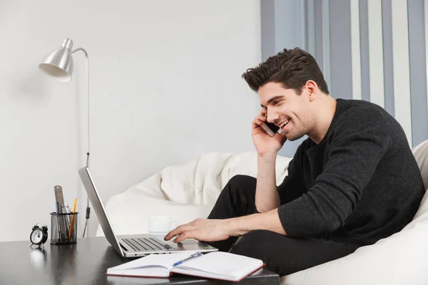 Przystojny młody człowiek w domu w pomieszczeniu przy pomocy laptop rachmistrz rozmowa przy zdejmowany telefon. — Zdjęcie stockowe