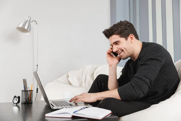 Beau jeune homme à la maison à l'intérieur en utilisant un ordinateur portable parlant par téléphone mobile . — Photo