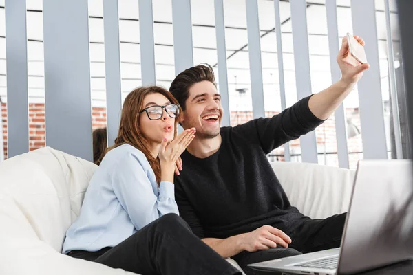 Szczęśliwa Młoda para kochająca w domu w pomieszczeniu za pomocą komputera przenośnego zrobić selfie przez telefon. — Zdjęcie stockowe
