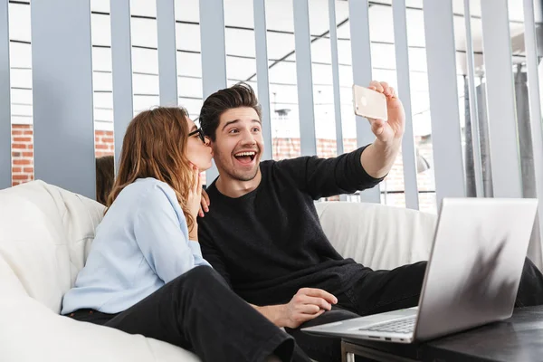 Happy young loving couple at home indoors using laptop computer make a selfie by phone. — Stock Photo, Image