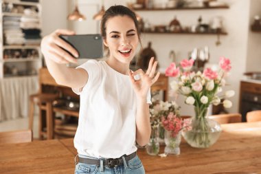 Beyaz tişörtlü güzel genç öğrenci kız kapalı mekanlarda poz vererek tamam jesti göstererek cep telefonuyla selfie çekti.