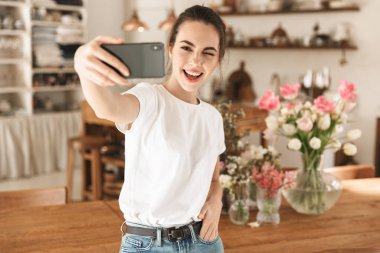 Beyaz tişörtlü güzel genç öğrenci kız kapalı mekanlarda poz vererek tamam jesti göstererek cep telefonuyla selfie çekti.