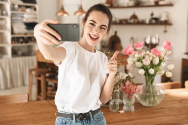 Beyaz tişörtlü güzel genç öğrenci kız kapalı mekanlarda poz vererek tamam jesti göstererek cep telefonuyla selfie çekti.