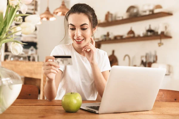 Menina estudante bonita com maçã dentro de casa usando computador portátil segurando cartão de crédito . — Fotografia de Stock